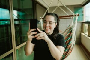 Woman smiling while taking a selfie with a smartphone on a colorful balcony with a hammock.